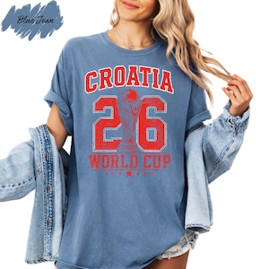 A woman wearing a blue t-shirt with the text "CROATIA 26 WORLD CUP" printed on it, along with a trophy icon. She is also wearing a denim jacket and has her hair styled in loose waves.