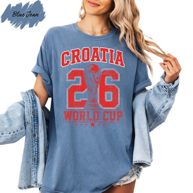A woman wearing a blue t-shirt with the text "CROATIA 26 WORLD CUP" printed on it, along with a trophy icon. She is also wearing a denim jacket and has her hair styled in loose waves.