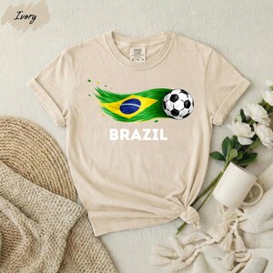 A beige t-shirt with a graphic of a soccer ball and the word "BRAZIL" printed on it, along with a Brazilian flag design. The t-shirt is placed on a white surface, and there are some flowers and a mug nearby.
