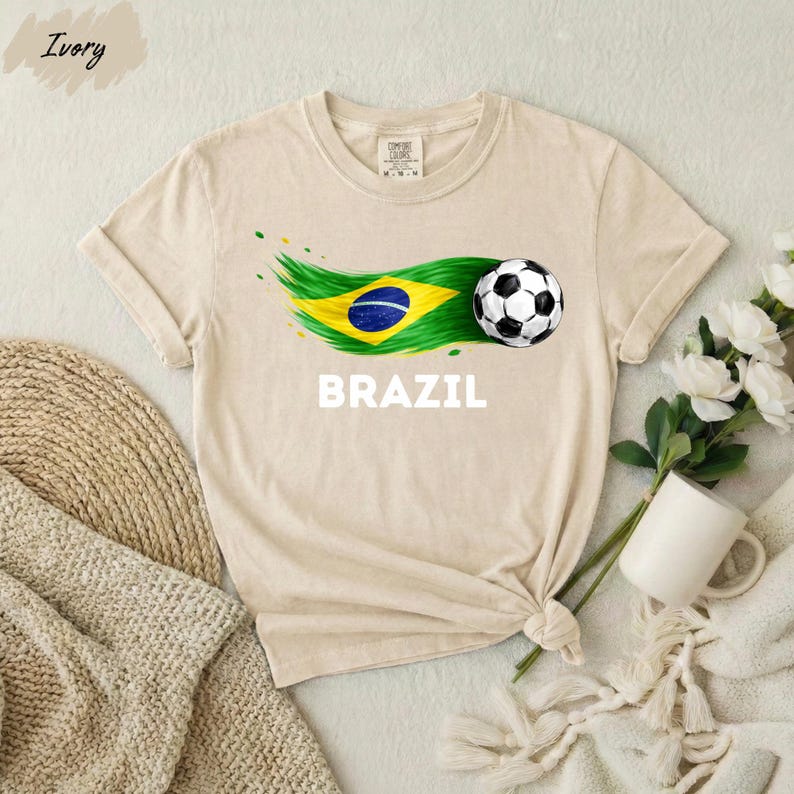 A beige t-shirt with a graphic of a soccer ball and the word "BRAZIL" printed on it, along with a Brazilian flag design. The t-shirt is placed on a white surface, and there are some flowers and a mug nearby.