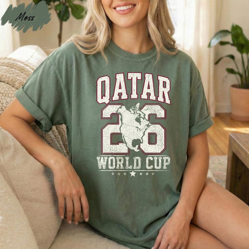 A woman wearing a green t-shirt with the text "QATAR 26 WORLD CUP" printed on it, sitting on a couch.