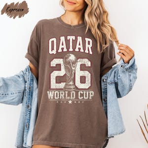 A woman wearing a brown t-shirt with the text "QATAR 26 WORLD CUP" printed on it, along with a trophy icon. She is also wearing a denim jacket and has her arms outstretched.