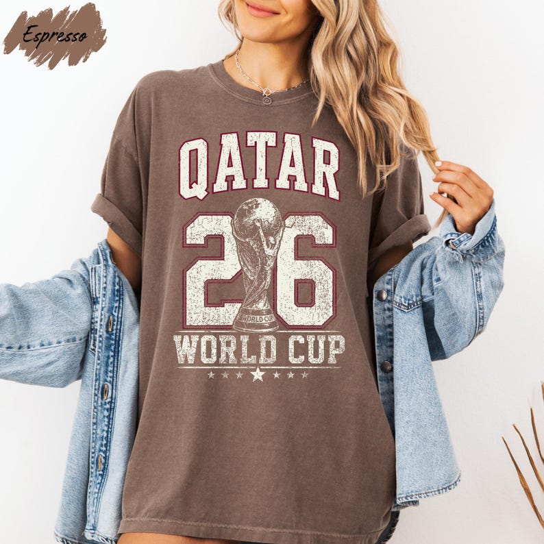 A woman wearing a brown t-shirt with the text "QATAR 26 WORLD CUP" printed on it, along with a trophy icon. She is also wearing a denim jacket and has her arms outstretched.