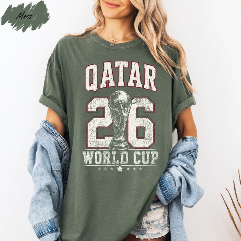 A woman wearing a green t-shirt with the text "QATAR 26 WORLD CUP" printed on it, along with a trophy icon. She is also wearing a denim jacket and has her hair styled in loose waves.