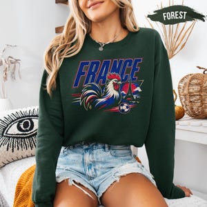 A woman wearing a green sweatshirt with a graphic of a rooster and the word "FRANCE" printed on it, along with denim shorts.