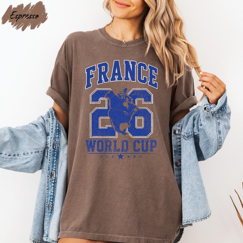 A woman wearing a brown t-shirt with the text "FRANCE 26 WORLD CUP" printed on it, along with a graphic of a globe. She is also wearing a denim jacket and has her arms outstretched.
