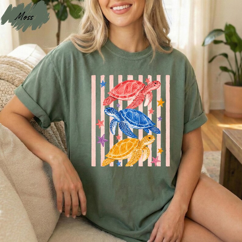 A woman wearing a green t-shirt with a colorful graphic of three sea turtles and stars on it, sitting on a couch.