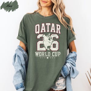 A woman wearing a green t-shirt with the text "QATAR 26 WORLD CUP" printed on it, along with a graphic of a globe. She is also wearing a denim jacket and denim shorts.