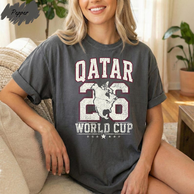 A woman wearing a grey t-shirt with the text "QATAR 26 WORLD CUP" printed on it, sitting on a couch.