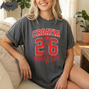 A woman wearing a grey t-shirt with the text "Croatia 26 WORLD CUP" printed on it, sitting on a couch.
