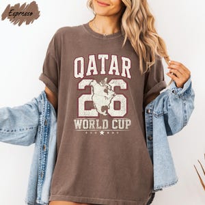 A woman wearing a brown t-shirt with the text "QATAR 26 WORLD CUP" printed on it, along with a graphic of a map. She is also wearing a denim jacket and has her arms outstretched.