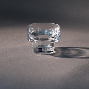 Puede incluir: Un portavelas de vidrio transparente con una base cilíndrica y una parte superior circular más ancha. El vidrio tiene un ligero tinte azul y proyecta una sombra sobre la superficie gris. Una pequeña etiqueta es visible en la base.