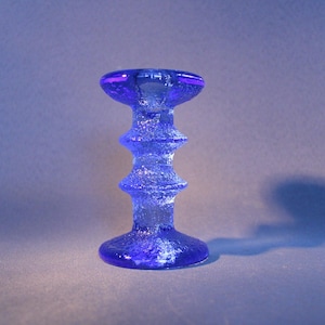 Puede incluir: Un candelero de vidrio azul cobalto con una superficie texturizada. El candelero tiene una base redonda, un tallo escalonado y una parte superior circular. El objeto mide aproximadamente 15 cm de altura.