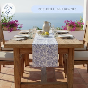 May include: A wooden dining table set for a meal, featuring a blue and white Delft table runner. The table is set with plates, glasses, and a ceramic pitcher. The text "BLUE DELFT TABLE RUNNER" is displayed above the table.