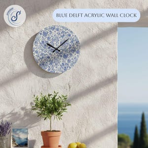 May include: A round, blue and white acrylic wall clock with a Delft floral pattern. The clock has black hands and the text "BLUE DELFT ACRYLIC WALL CLOCK" is above it. The clock is mounted on a textured white wall.