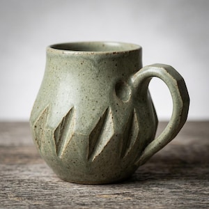 Puede incluir: Taza de cerámica verde salvia hecha a mano, moteada, con un diseño geométrico tallado. La taza tiene un cuerpo redondeado, un asa facetada y una capacidad de aproximadamente 350 ml.