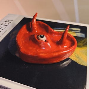 May include: A heart-shaped, bright red ceramic dish with two horns and a single eye. The glossy dish is displayed on a stack of books, with artist names visible.