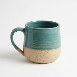 May include: A handmade ceramic mug with a teal glazed top and a speckled beige bottom. The mug has a rounded shape with a large handle. The mug is perfect for coffee, tea, or any hot beverage.
