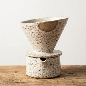 May include: A speckled ceramic pour-over coffee maker set. The set includes a cone-shaped dripper with a brown accent and a matching base with a spout. The coffee maker is light beige with brown speckles.