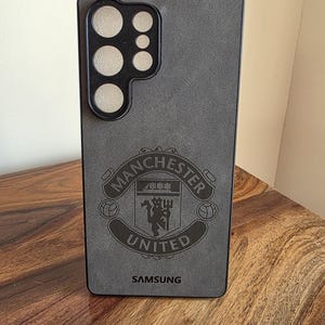 May include: Grey smartphone case with the Manchester United emblem and the word "SAMSUNG" at the base. The case has a camera aperture and a textured surface. Designed for a mobile phone.