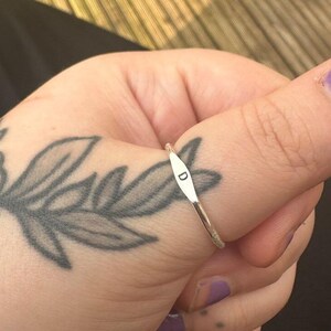 May include: A silver ring with a slender band and a small, elongated plate engraved with the letter "D". The ring is worn on a thumb with purple nail polish and a floral tattoo.
