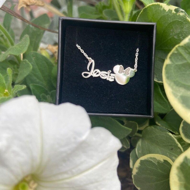 May include: A silver necklace in a black box. The necklace spells out the name "Josie" in cursive script, with a Mickey Mouse head charm. The necklace is displayed against a backdrop of green foliage and a white flower.