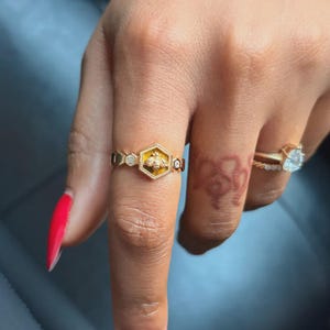 May include: A gold ring with a hexagonal bee design, featuring a yellow enamel background and small gemstones. Another ring with a large, clear gemstone is also visible. The rings are worn on a finger.