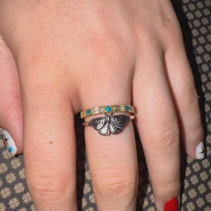 May include: Two rings are displayed on a finger. The bottom ring is silver with a butterfly design. The top ring is gold with clear and turquoise-colored stones. The background is a patterned fabric.