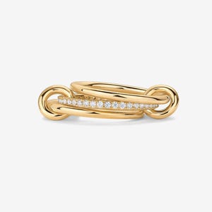 May include: A gold ring set with three interlocking bands. One band is set with small, clear gemstones. The design features rounded, looped accents at each end, creating a unique and elegant look. The rings are displayed against a plain background.