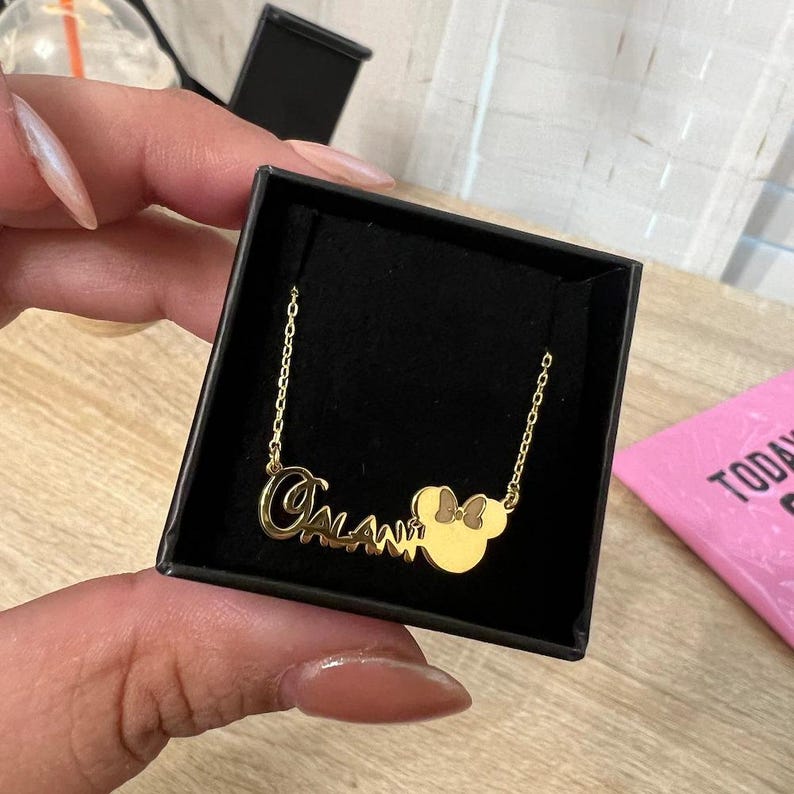 May include: A gold-colored necklace with the name "Calani" in cursive script and a Minnie Mouse head pendant. The necklace is displayed in a black box, with a pink card visible in the background.