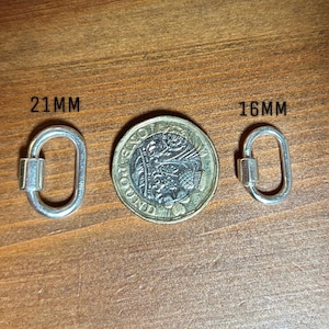 Puede incluir: Dos cierres de mosquetón ovalados plateados, uno de 21 mm y otro de 16 mm, se muestran junto a una moneda. Los cierres están sobre una superficie de madera. Los cierres se utilizan para la fabricación de joyas o manualidades.