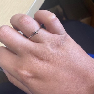 May include: A silver ring with a small, geometric design. The ring is on a finger, and the design appears to be a stylized lightning bolt or zig-zag shape. The ring is delicate and minimalist.