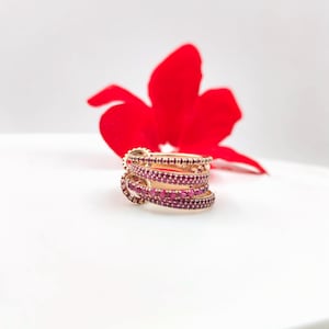 Könnte beinhalten: Ein Stapel goldfarbener Ringe mit mehreren Bändern, die jeweils mit kleinen, leuchtend roten Edelsteinen besetzt sind. Die Ringe sind vor einem weichen, weißen Hintergrund platziert, mit einer großen, leuchtend roten Blume im Hintergrund.