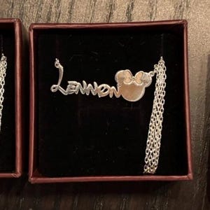May include: A silver necklace with the name "Lennon" in a cursive font, connected to a Minnie Mouse head pendant with a bow. The necklace is displayed in a small, dark red box with a black interior.