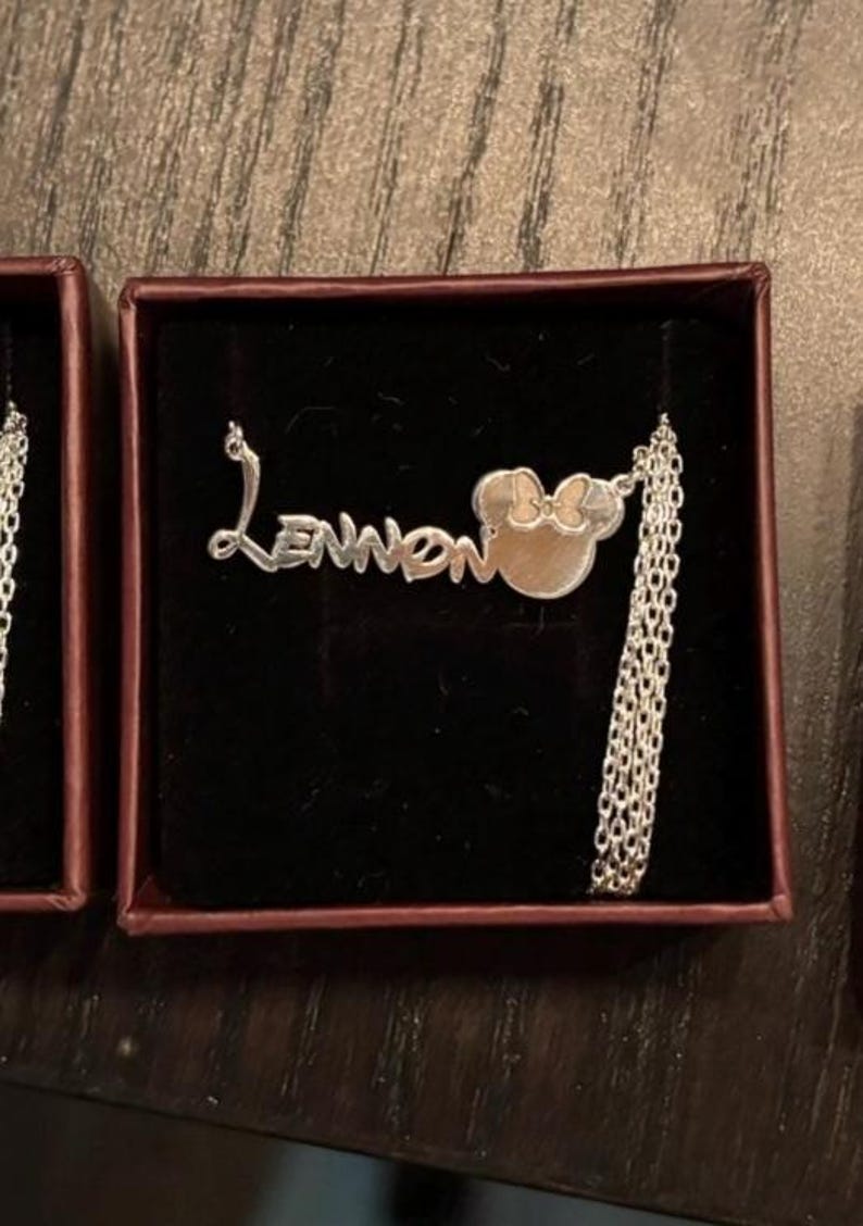 May include: A silver necklace with the name "Lennon" in a cursive font, connected to a Minnie Mouse head pendant with a bow. The necklace is displayed in a small, dark red box with a black interior.