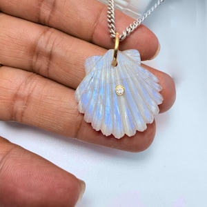 May include: A pendant shaped like a scallop shell carved from iridescent moonstone with a blue sheen. The shell features a small, round, clear gemstone set in a gold bezel at the centre and hangs from a silver chain by a gold bail.