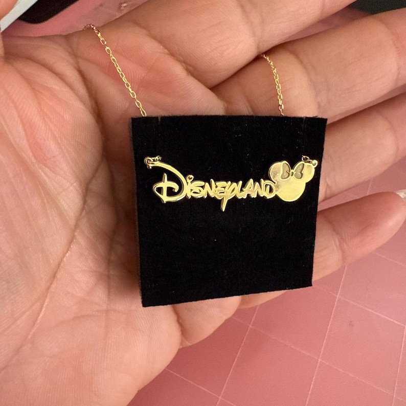 May include: Gold-colored necklace featuring the Disneyland logo and a Minnie Mouse silhouette. The pendant is displayed on a black velvet card. The chain is delicate and the overall design is elegant.