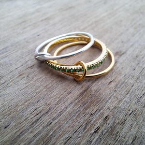 May include: Three rings, one silver and two gold, are interlinked. One gold ring is embellished with small green gemstones. The rings are set against a rustic, wooden surface, creating a contrast of textures and colours. The design suggests a theme of interconnectedness.