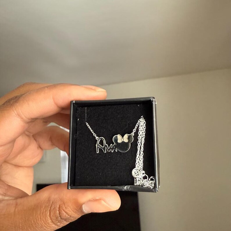 May include: A silver necklace with the name "Ava" and a Minnie Mouse head pendant with a bow. The necklace is displayed in a black box.