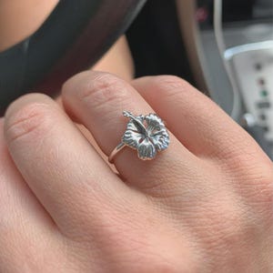 Può includere: Un anello d'argento con un design dettagliato di un fiore di ibisco. La fascia dell'anello è sottile e il fiore è il punto focale, che mostra dettagli intricati dei petali. L'anello è indossato su un dito, evidenziando i gioielli floreali.