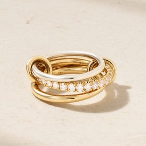 Op de afbeelding: Een stapel gouden ringen met een zilveren band en een band met kleine, heldere edelstenen. De ringen zijn in elkaar verweven, waardoor een gelaagd effect ontstaat. De ringen zijn geplaatst tegen een neutrale achtergrond.