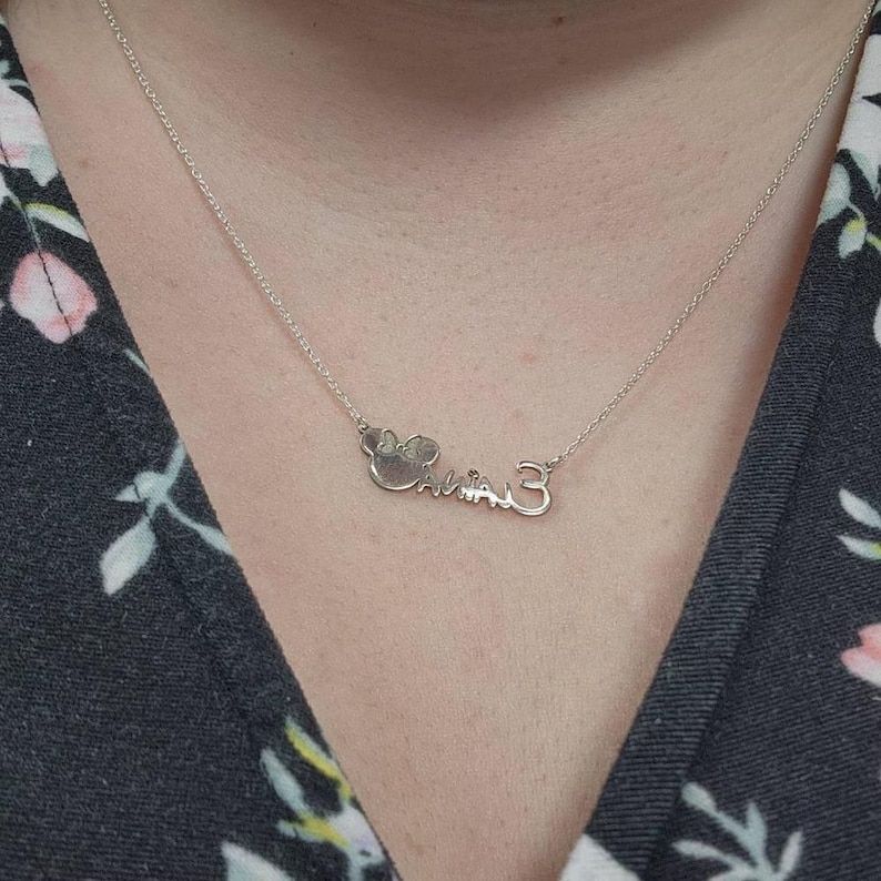 May include: A silver necklace with a pendant spelling out the name "Calina" in a cursive font, with a bow-shaped design at the beginning and the number "3" at the end. The necklace is on a delicate chain.