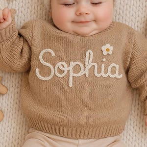 May include: A baby wearing a beige sweater with the name "Sophia" embroidered in white, a white knit hat with two pom-poms, and beige pants. Wooden toys, including a horse and giraffe, are placed around the baby on a cream-colored knitted blanket.