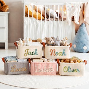 May include: A set of six corduroy storage baskets in various colors, each labeled with a name in embroidered script: Jack, Noah, Lucas, Isabella, and Oliver. The baskets are filled with baby items and toys, with leather handles.