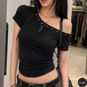 Puede incluir: Top negro asimétrico de manga corta con tachuelas plateadas en el escote y el hombro. El top tiene un diseño asimétrico y se combina con un collar y un cinturón. El top está hecho de un material suave.