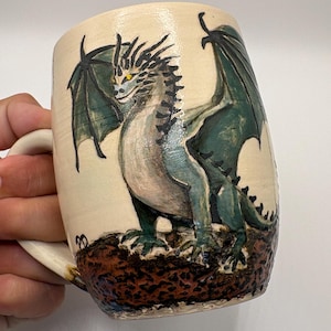 May include: A ceramic mug with a cream-coloured base featuring a hand-painted green dragon with yellow eyes and dark wings. The dragon stands on a textured, dark brown rocky surface at the bottom of the mug. The handle is attached to the left side.