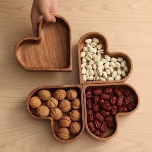 May include: Wooden heart-shaped serving dishes, one held by a hand. Three dishes are filled with pistachios, walnuts, and red dates. The dishes are made of a light brown wood and sit on a light wood surface.
