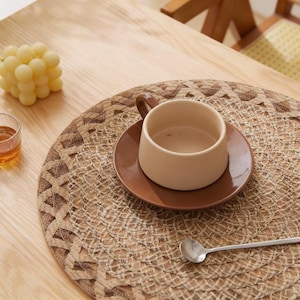 Peut inclure: Une tasse et une soucoupe en céramique beige sur un dessous de plat tissé. Une petite cuillère en argent repose sur le dessous de plat. Un verre de liquide et une grappe de raisins sont également visibles sur la table en bois.