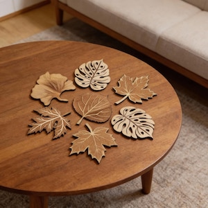 May include: Seven wooden leaf-shaped coasters in different designs, such as maple and monstera leaves, are displayed on a round, dark wood coffee table. The coasters are light brown with detailed veining. The table is made of dark wood.