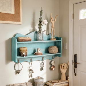 May include: A turquoise wooden wall shelf with a decorative wavy edge, displaying a candle, wooden figurine, and twine. Metal hooks below hold keys and a ring. A corkboard and a white door are visible in the background.